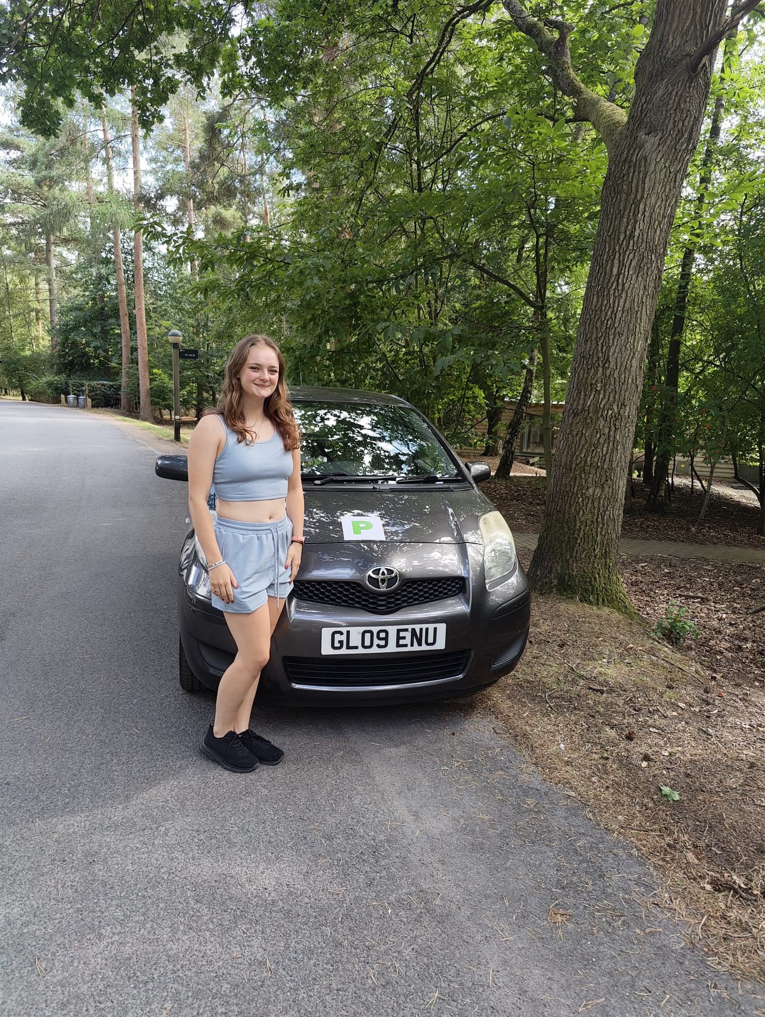 Jess standing beside her new grey Toyota Yaris with green P plates on a woodland holiday-park road