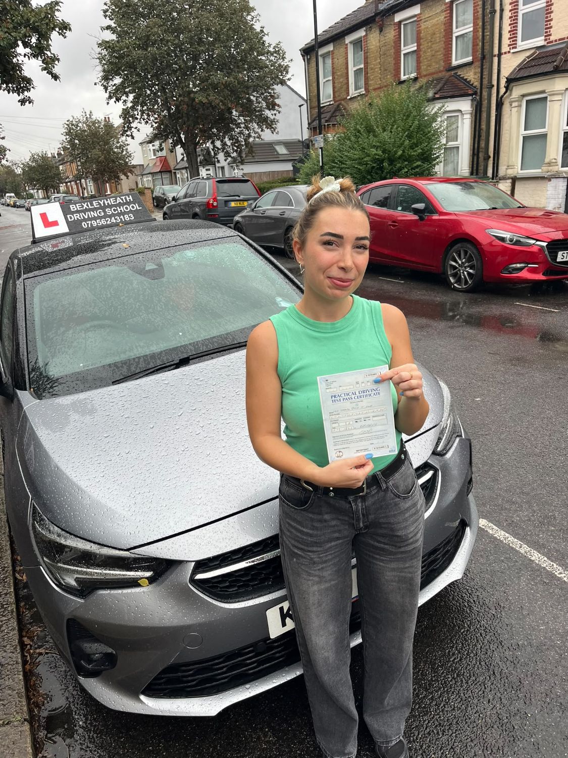 Bella standing in front of the Bexleyheath Driving School car in the rain holding her practical driving test pass certificate
